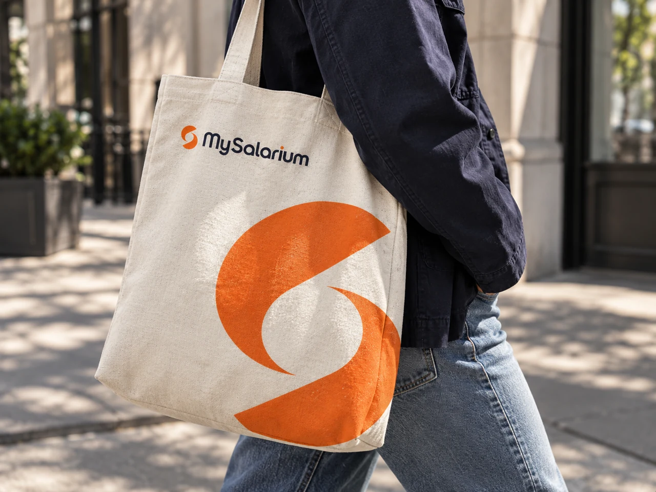 Cream canvas tote with the MySalarium signature and orange symbol carried on a city sidewalk